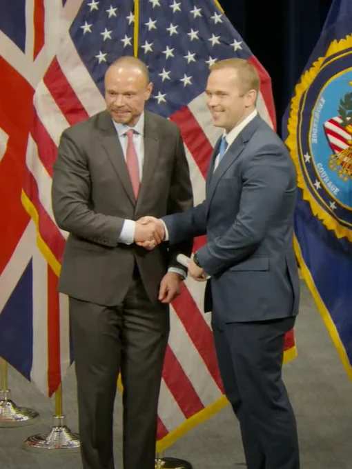 Two individuals shaking hands in front of multiple United States and United Kingdom flags, symbolizing a diplomatic or formal event.