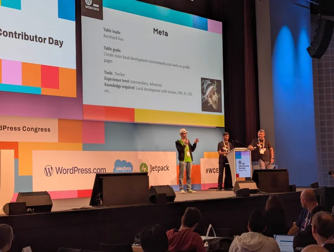 Bernhard from Syde stands on stage at WordCamp Europe Contributor Day 2025, introducing the Meta table as table lead. The large screen behind him shows table details, including goals and tools. Two other contributors are on stage alongside him, with attendees watching from the audience.