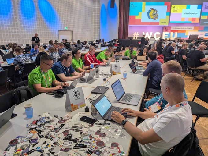 A large group of contributors at WordCamp Europe Contributor Day 2025 are working together at long tables, laptops open and focused on WordPress-related tasks. In the foreground, a pile of WordPress stickers is spread across the table. Several Syde team members in green shirts are visible, actively participating in the collaborative atmosphere.