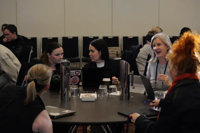 Five women sit at a round table with computers open in front of them. They're talking with each other. 