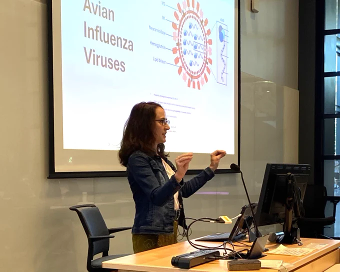 Woman with long brown hair and glasses gestures while standing in front of a projected image with text reading: Avian Influenza Viruses