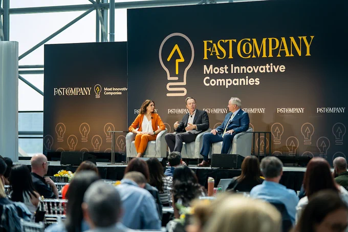 Three individuals representing Texas A&M are seated on stage at a Fast Company event discussing the Most Innovative Companies. They are engaged in a conversation in front of a branded backdrop with an audience in the foreground.