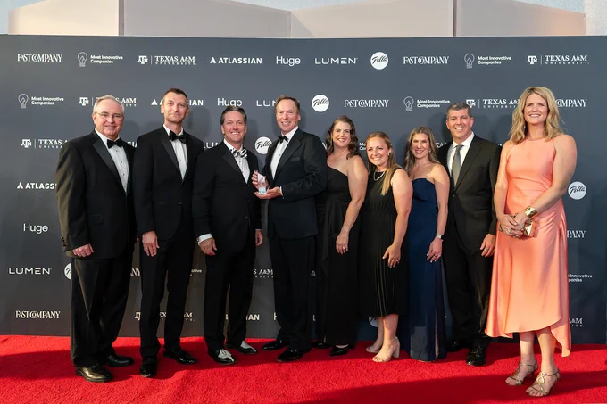 Group of nine individuals posing together on a red carpet at a Texas A&M event, holding an award.