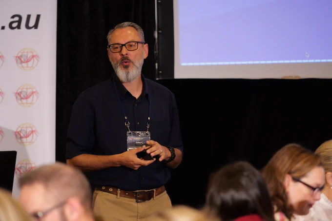A man in a blue shirt with a beard and glasses holds his hands in front of him while speaking to a group of people. 