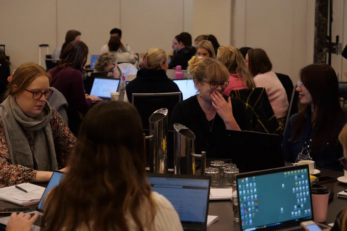 A group of women with laptops on their tables speak in groups. 