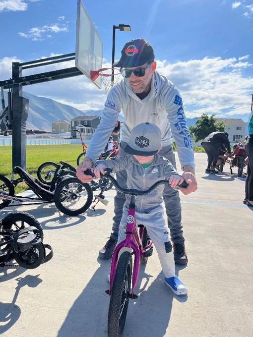a man stands behind a bike with a boy on it, they both hold the handlebars. there are other accessible vehicles behind them and a basketball hoop.