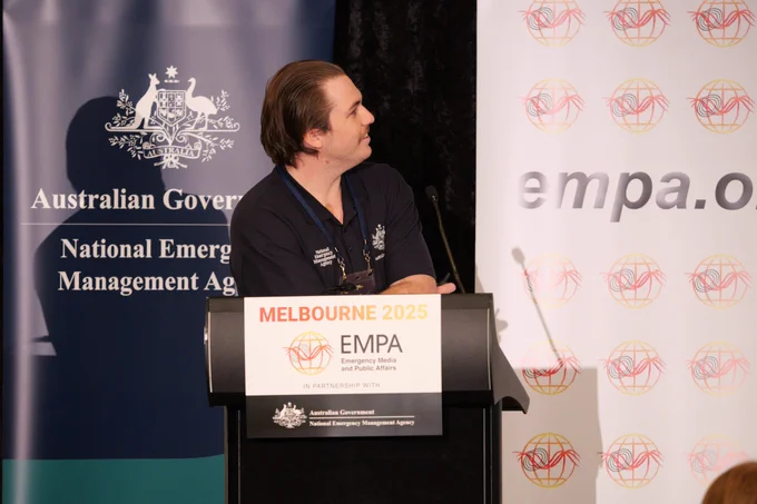 A man in a blue tshirt with brown hair looks to his left. There are two banners behind him, NEMA and EMPA. 