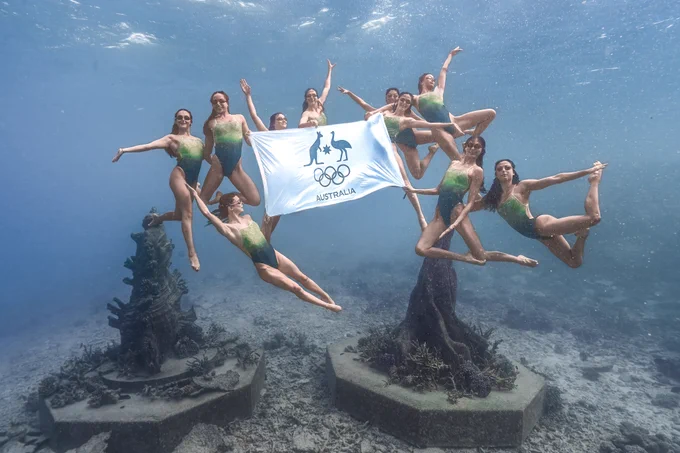 Australia's artistic swimming team at the Museum of Underwater Art. Each team member is holding different poses as they hold the Australian Olympic Committee flag.