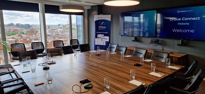 An empty meeting room with a large wooden table set with glasses, pens, and notepads. The room features floor-to-ceiling windows overlooking a cityscape, a banner for OrbusInfinity, and screens displaying 'Orbus Connect Victoria Welcome.' 