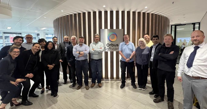 A group of people standing in front of a wooden panel wall featuring the Orbus Software logo in a modern office setting 