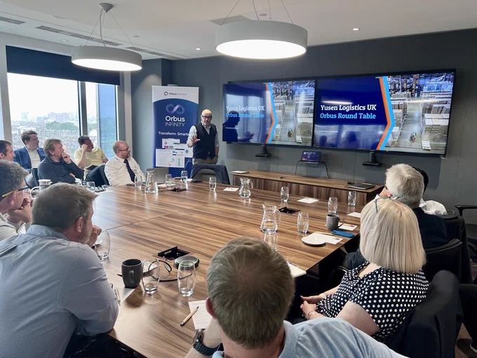 A meeting room with attendees seated around a large wooden table, listening to a presenter standing near screens displaying the title 'Yusen Logistics UK Orbus Round Table.' A banner for OrbusInfinity is visible in the background. 