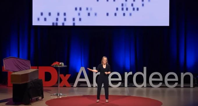 Dr Jennifer Riley on stage, next to a coffin and a round table. TEDx Aberdeen displayed at the back.