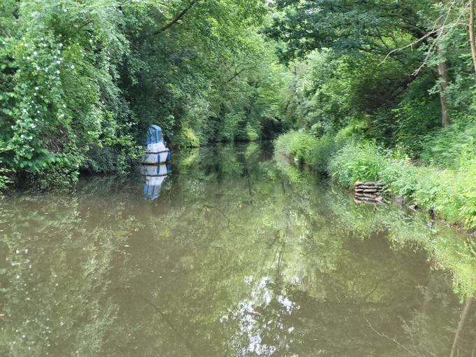 A canal with trees and foliage on the embankment, with a blue and white canal boat parked on the left hand side