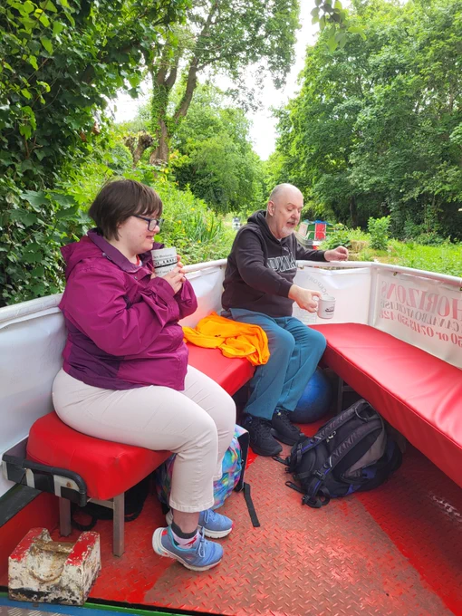 A man and a woman drinking from mugs sat at the front of a canal boat. 