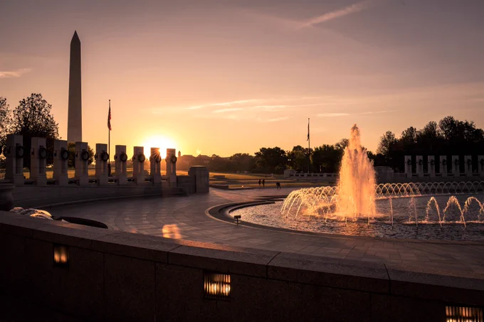 The World War II Memorial in Washington, D.C., at sunrise. The sun rises just beyond the pillars and flagpole, casting a golden glow across the plaza. A large fountain is in the foreground and the Washington Monument stands tall in the background. 
