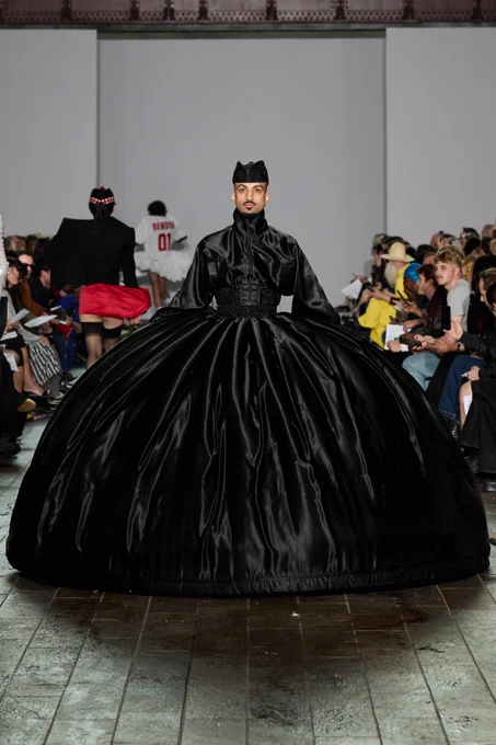 A model wears a dramatic full-length black gown with an exaggerated voluminous skirt and structured shoulders, walking down the runway.