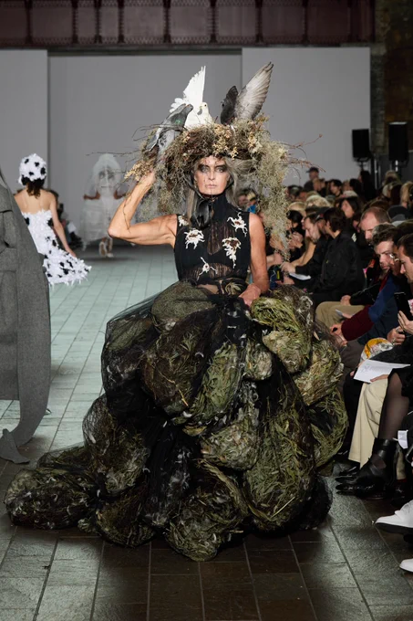 A model wears a textured, earth-toned dress with a large bird's nest headpiece featuring artificial birds and dried foliage.