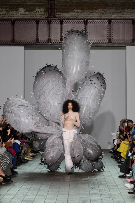 A model walks in a sheer, pale outfit surrounded by massive, grey, inflatable feathered forms that extend from their back, creating a cocoon-like structure.