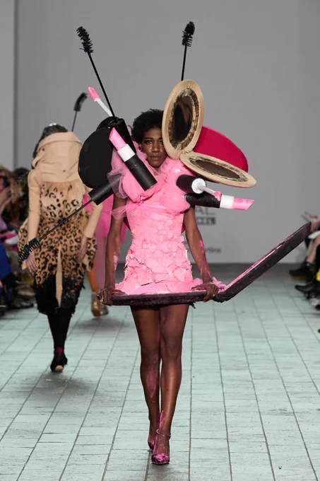 A model walks the runway in a sculptural pink dress surrounded by oversized props resembling makeup items like mascara wands, lipsticks, and powder compacts.
