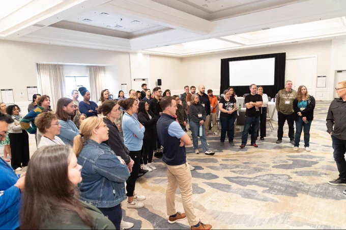 
Crowd of people in a white room surrounding the person speaking (right)