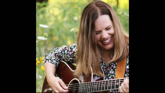 Joy Zimmerman playing an acoustic guitar and smiling, surrounded by greenery.
