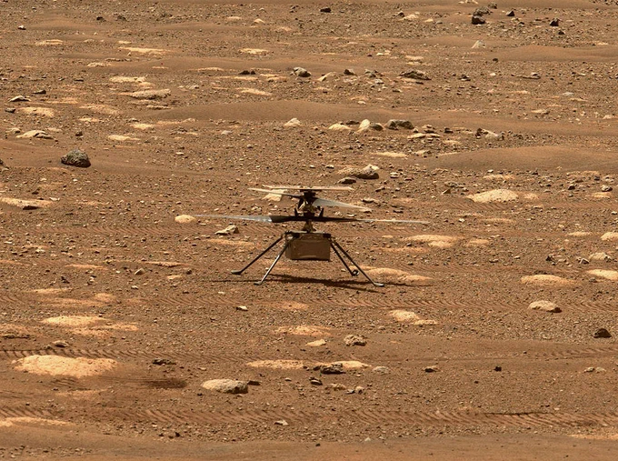 The Ingenuity Mars Helicopter is shown in a very rocky and sandy area of Mars. The silver and gray rotorcraft is stationary, with four rotor blades stacked on top of its boxy body as it stands on the reddish-hued surface.