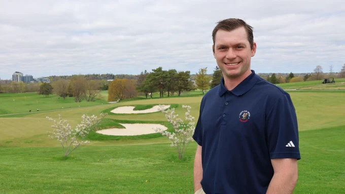 A man with brown hair wearing a navy golf shirt stands smiling with a golf course and buildings in the background.