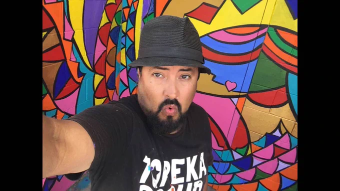 Robert Tapley Bustamante wearing a fedora and a 'Topeka' T-shirt taking a selfie in front of a colorful, abstract mural.