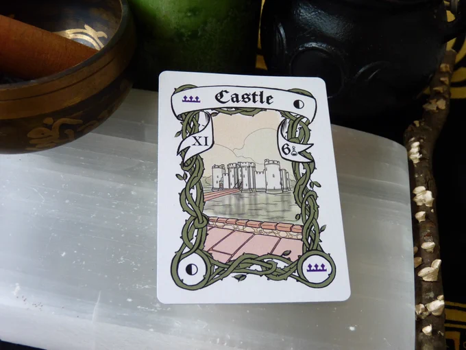 The Quarter Moon Castle card has a quarter moon in 2 corners and a purple crown in the other two, with green border vines framing a view of a white and green castle from afar. It is surrounded by water with a long path, and we view it from a road.  In the banner there is the Roman Numeral XI on the left side and the Arabic number 6 on the right. The card is laying on a selenite slab, surrounded by a singing bowl, a green candle, a triple moon goddess cauldron, and a stick with lichen, all on a black and yellow cloth.