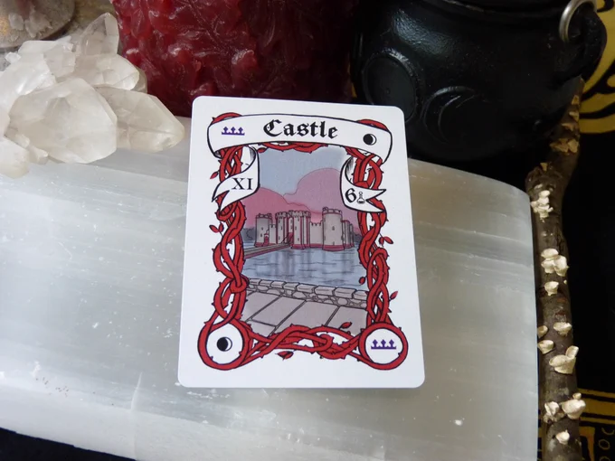 The Crescent Moon Castle card has a crescent moon in 2 corners and a purple crown in the other two, with red border vines framing a view of a grey and red castle from afar. It is surrounded by water with a long path, and we view it from a road.  In the banner there is the Roman Numeral XI on the left side and the Arabic number 6 on the right. The card is laying on a selenite slab, surrounded by a cluster of clear quartz, a red candle, a triple moon goddess cauldron, and a stick with lichen, all on a black and yellow cloth.
