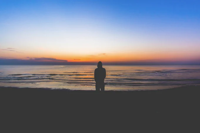 Photo of a silhouette of a person against dawn backdrop, at the sea. Grainy, dramatic image.