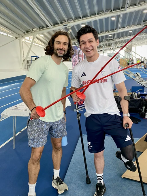 Two people holding plastic swords smile to the camera. They are in an athletics centre.