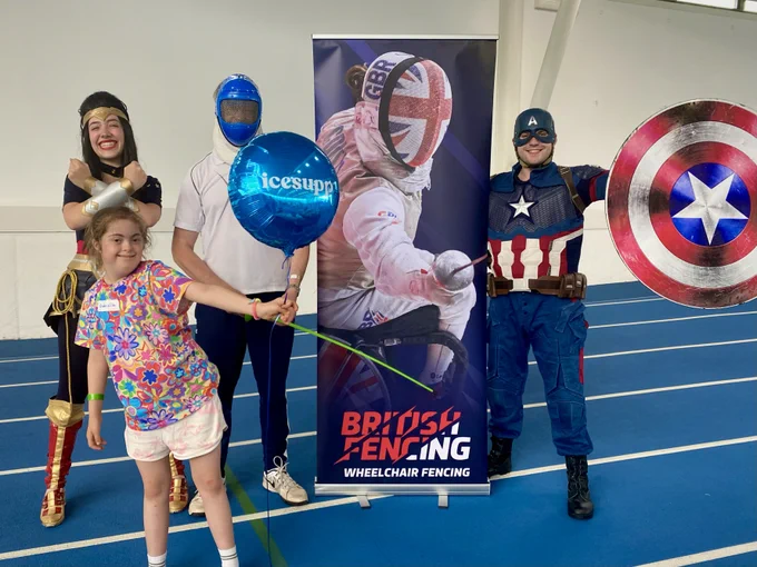 A photo of four people smile and pose to the camera. One is a child holding a balloon, one is wearing a Captain America costume, one is wearing a Wonder Woman costume, and the other is wearing a plastic fencing mask and holding a sword. They are next to a British Fencing wheelchair fencing banner.