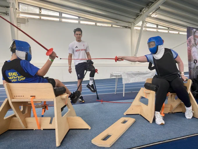 Two people are fencing on a wooden SwordSeat frame, wearing plastic masks and with plastic swords. Another person is refereeing.
