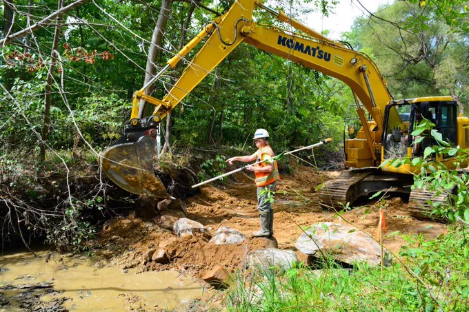 Environmental technician directing riprap placement with a Komatsu excavator during active streambank stabilization to prevent erosion along a muddy streambed.