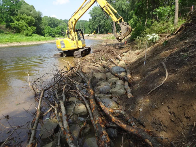 Excavator installing woody debris and rock toe protection along a steep streambank to prevent erosion and reinforce channel stability using natural materials.