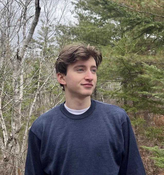 Darcy Eygun standing outdoors in front of trees, wearing a blue sweater. The image accompanies an announcement of his 3M National Student Fellowship award.