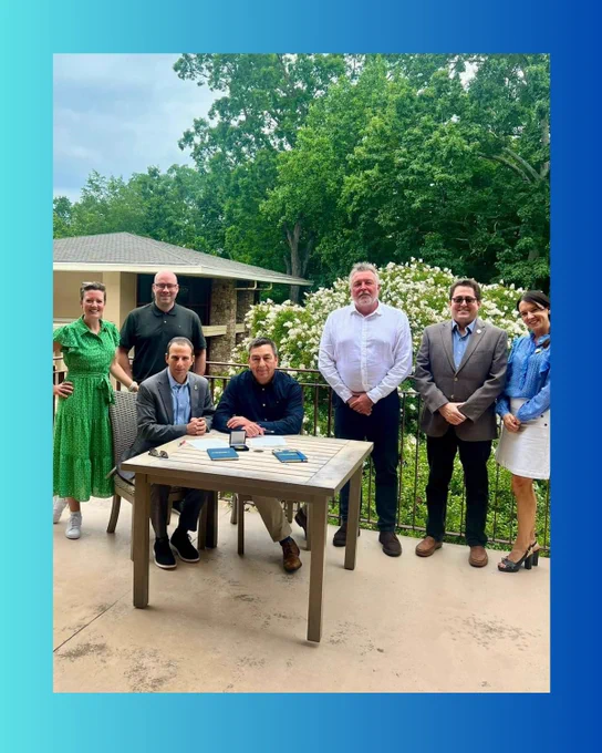 Childlight CEO Paul Stanfield and CRC CEO Greg Schiller sign the memorandum of understanding at a table outside. Behind them stand members of both teams. 