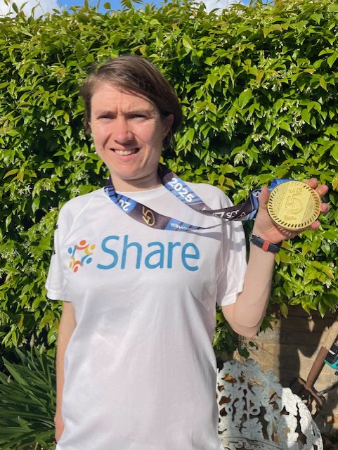 Georgia, a Share student, standing in front of a green bush wearing a Share running t-shirt and a gold medal around her neck. She is smiling and proudly holding up the medal.