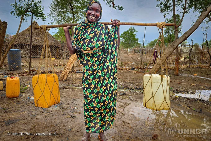 Photo description: Smiling girl in a green dress carrying two jerry cans of water hanging from a neck yoke.

Photo credit: UNHCR/Philip James Luduku