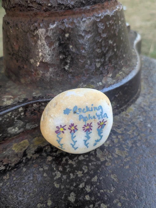 A pale pebble that has #RockingAphasia written on it with purple flowers underneath 