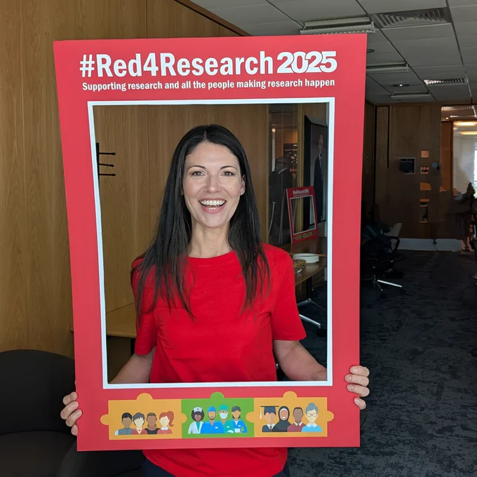 Person in a red shirt holding a large frame labeled "#Red4Research 2025" which supports research and all the people making research happen.