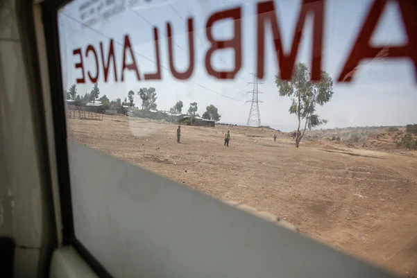 Image shows fields in Ethiopia through an ambulance window. There are people standing on the field and ambulance is written in red text across the window.