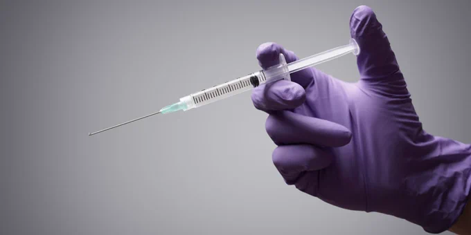 A hand wearing a purple medical latex glove holding a syringe ready to inject.