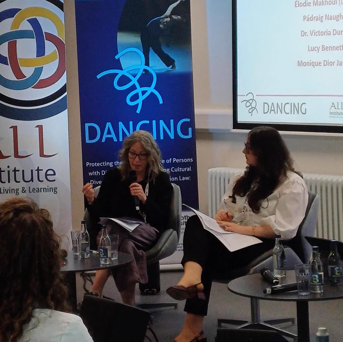 Photo of Victoria Durrer speaking into a microphone at the conference, with a backdrop of banners of contributing organisations.