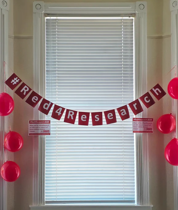 #Red4Research banner on display in the window of in the NI Clinical Trials Unit surrounded by red balloons.