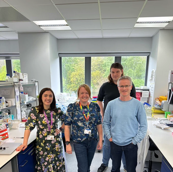 Dr Brian Conway, a male with short grey hair and glasses, stands in a laboratory with three members of his team. One tall male with dark brown hair, and two females, one with shoulder length brown hair and the other with light brown hair in a ponytail. There is science equipment visible on the desks either side of them. 