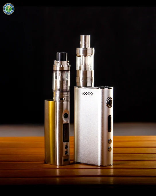 Two electronic cigarettes, one gold and one silver, standing upright on a wooden surface against a dark background.