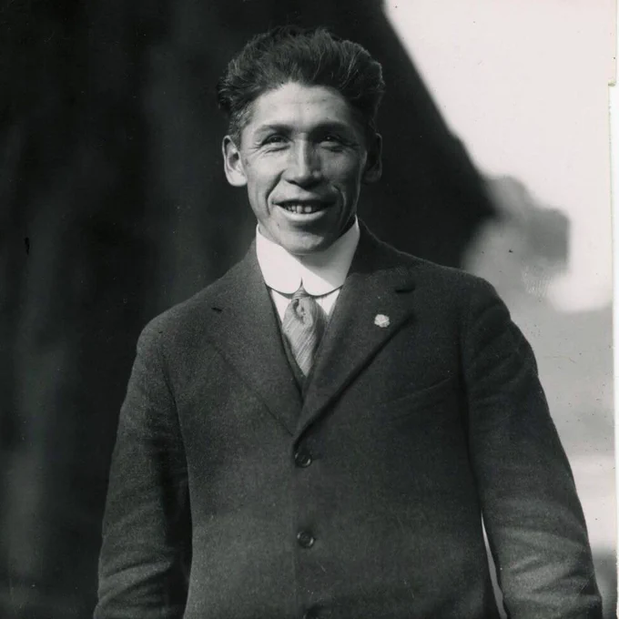 The black-and-white photo depicts Tom Longboat dressed in a formal suit with a high-collared shirt and tie. He is wearing a three-piece suit with a lapel pin on the left side. The background appears to be outdoors, with a dark, possibly tree-like shadow behind him. The image has a vintage feel, consistent with early 20th-century photography.