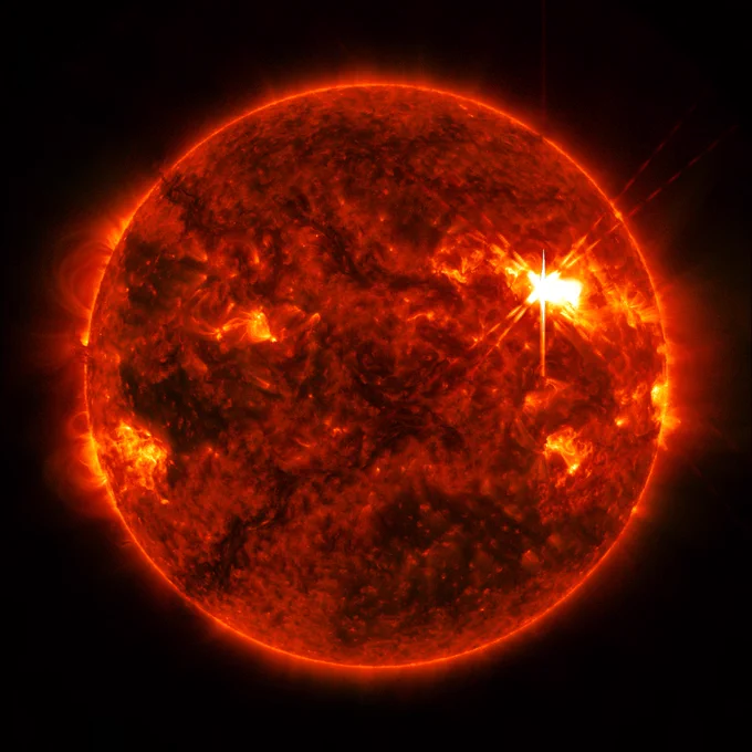 An extreme ultraviolet view of the Sun shows the bright flash of a solar flare on the Sun’s upper right side. The rest of the surface is mottled with darker and lighter regions, and faint loops of solar material can be seen extending off the Sun’s edges. 